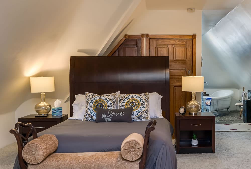 Dark wood sleigh bed, foot bench with tan cushions, two dark wood nightstands, two modern lamps and claw foot tub in background.