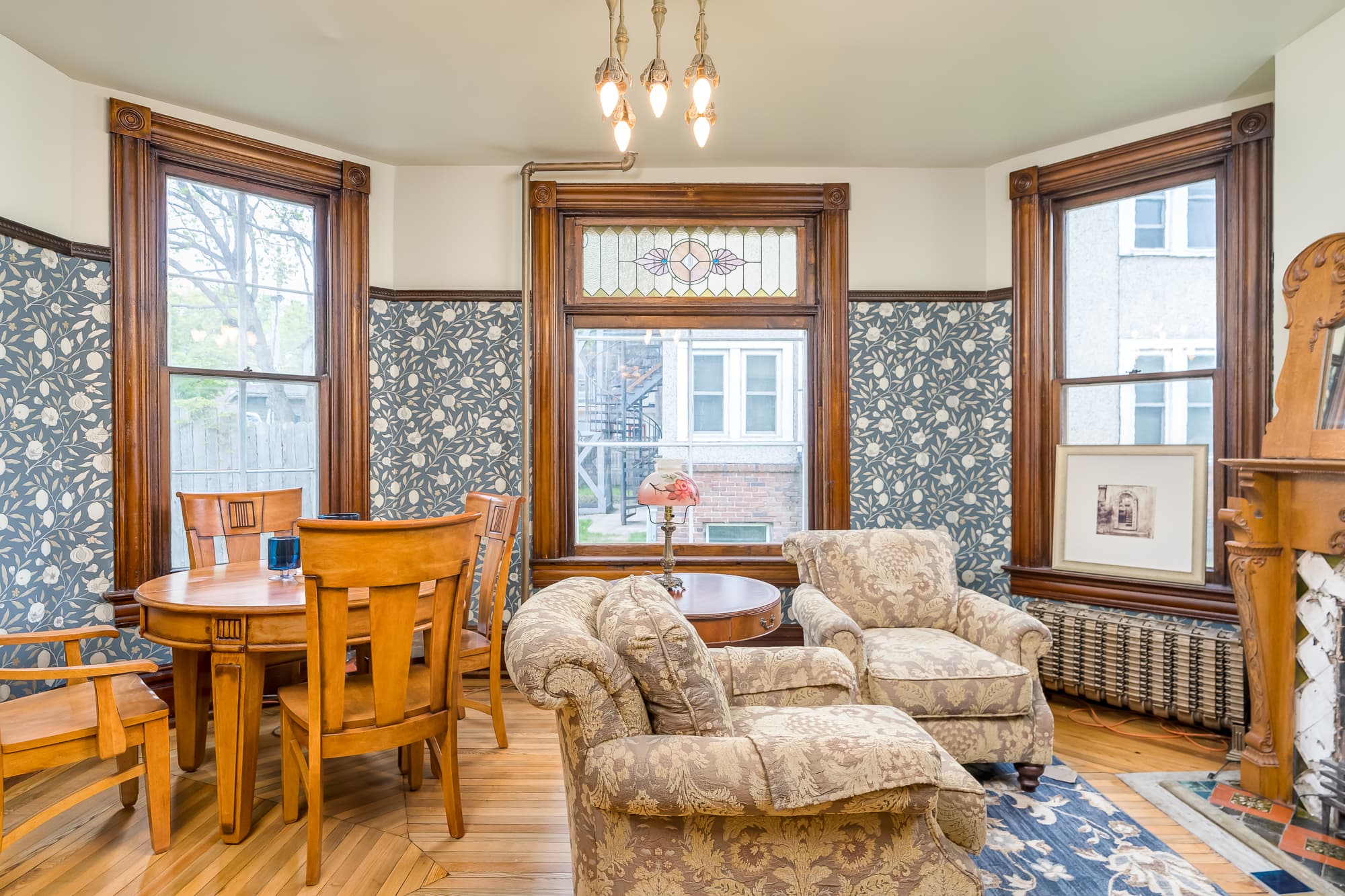 A cozy dining area features a wooden table and chairs beside floral wallpaper and large windows.