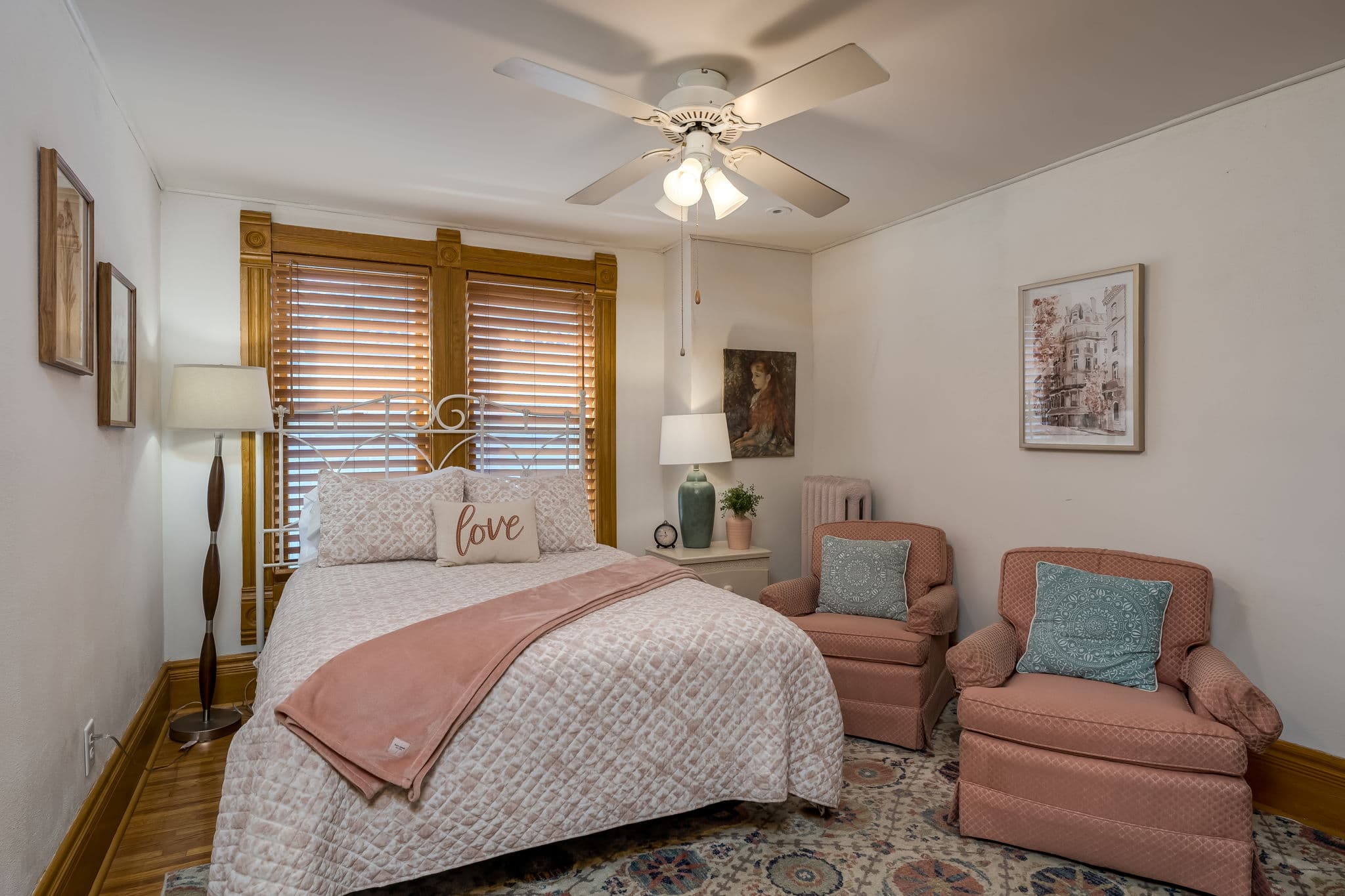 A cozy bedroom with a double bed, two pink armchairs, a ceiling fan, and warm wooden accents.