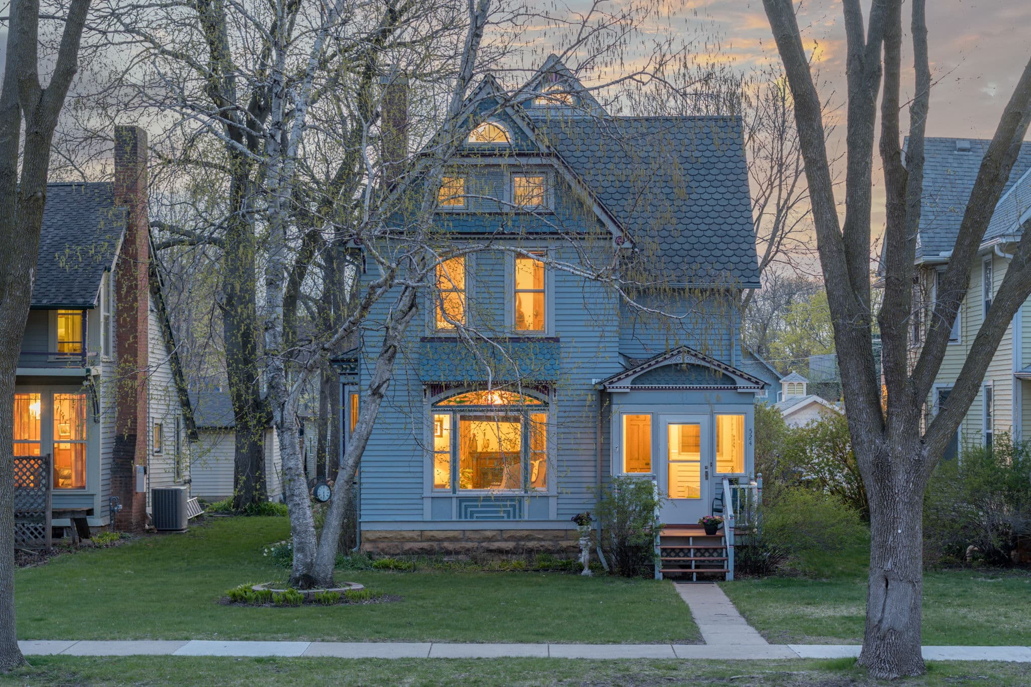 A blue Victorian house with glowing windows, surrounded by trees and a grassy lawn.