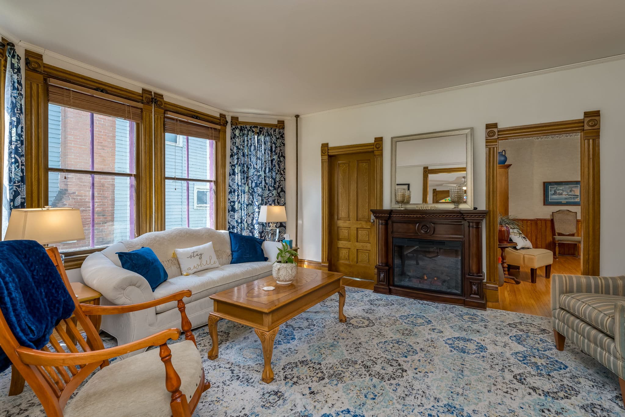 A cozy living room featuring a white sofa, wooden furniture, a decorative rug, and large windows with blue curtains.