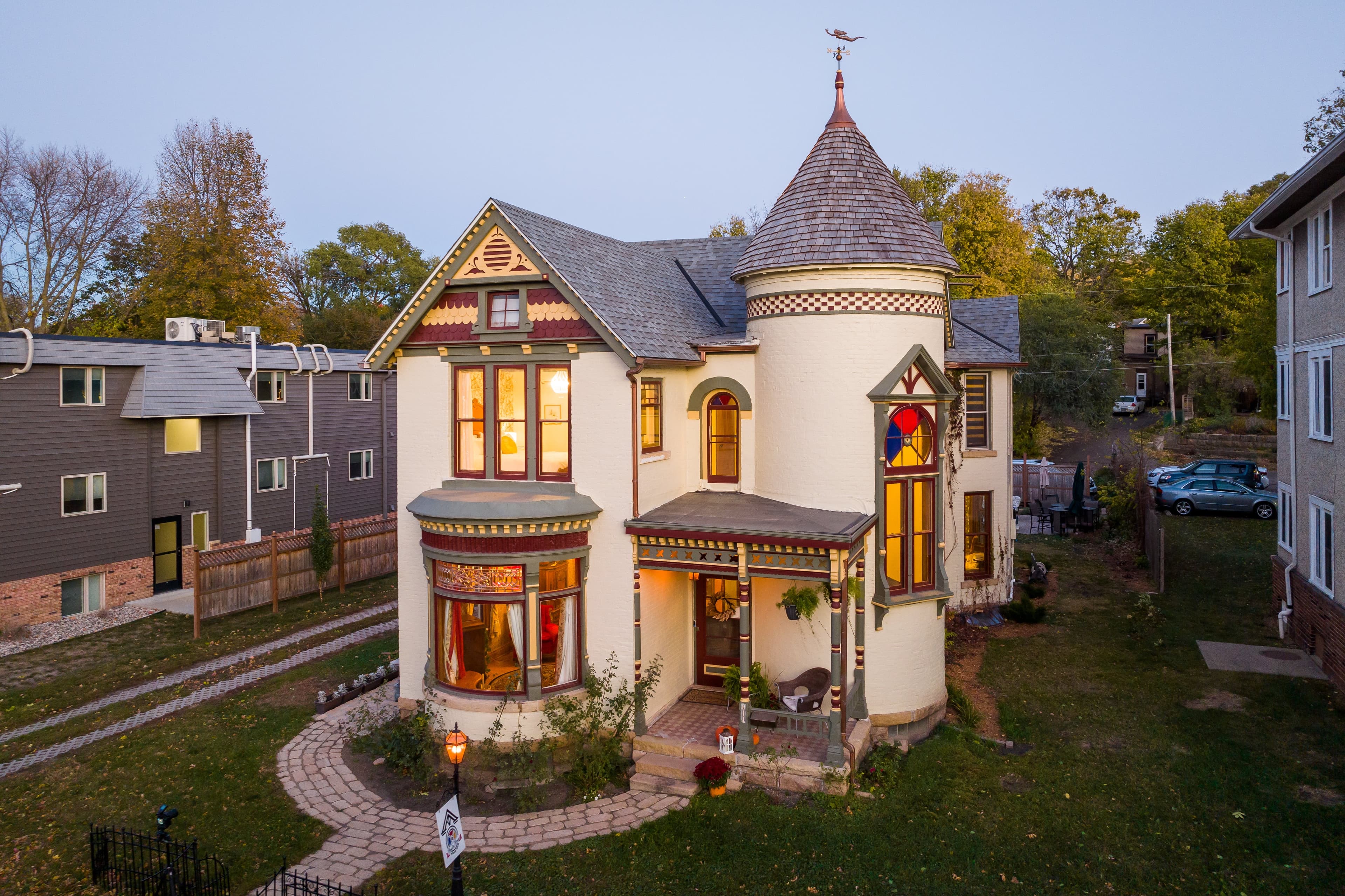 Victorian-style house with a turret and colorful stained glass windows, surrounded by landscaped gardens and other homes.