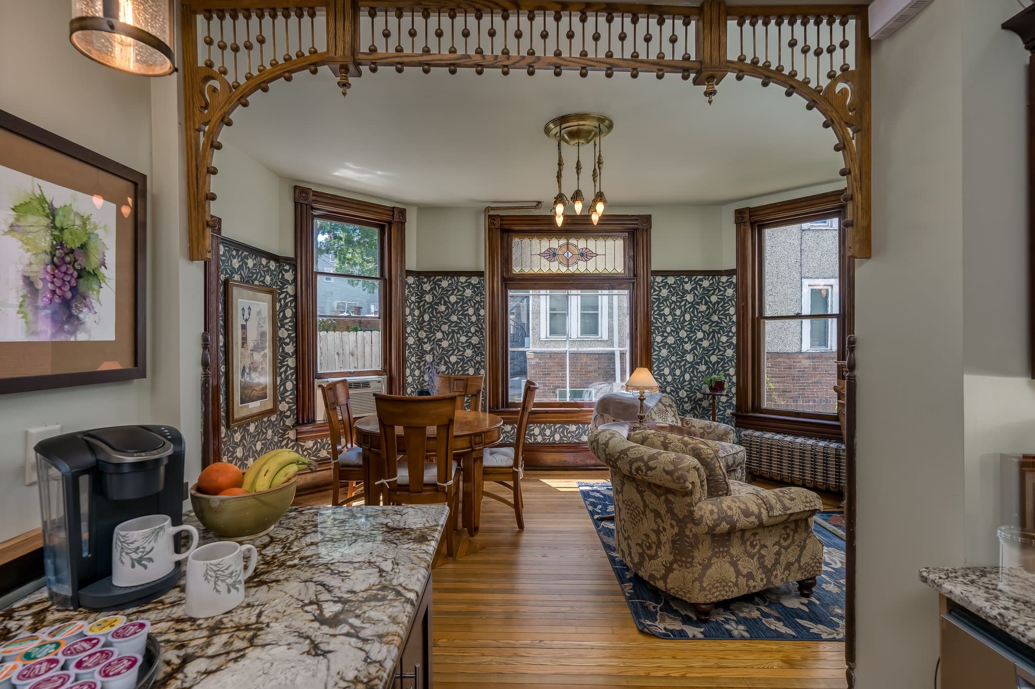 A cozy dining area with a wooden table, plush couch, and large windows, framed by decorative architecture and a countertop with a coffee maker and fruit.