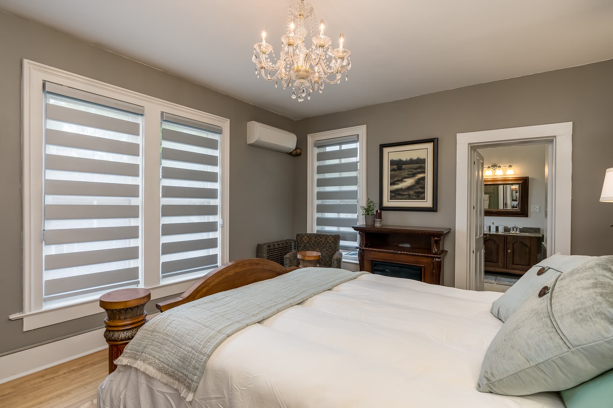 A cozy bedroom featuring a queen bed, chandelier, and large windows with striped shades.