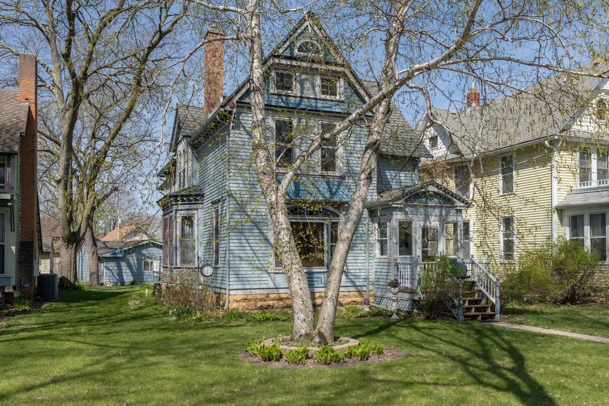 A blue Victorian house surrounded by trees and lush green lawn on a sunny day.