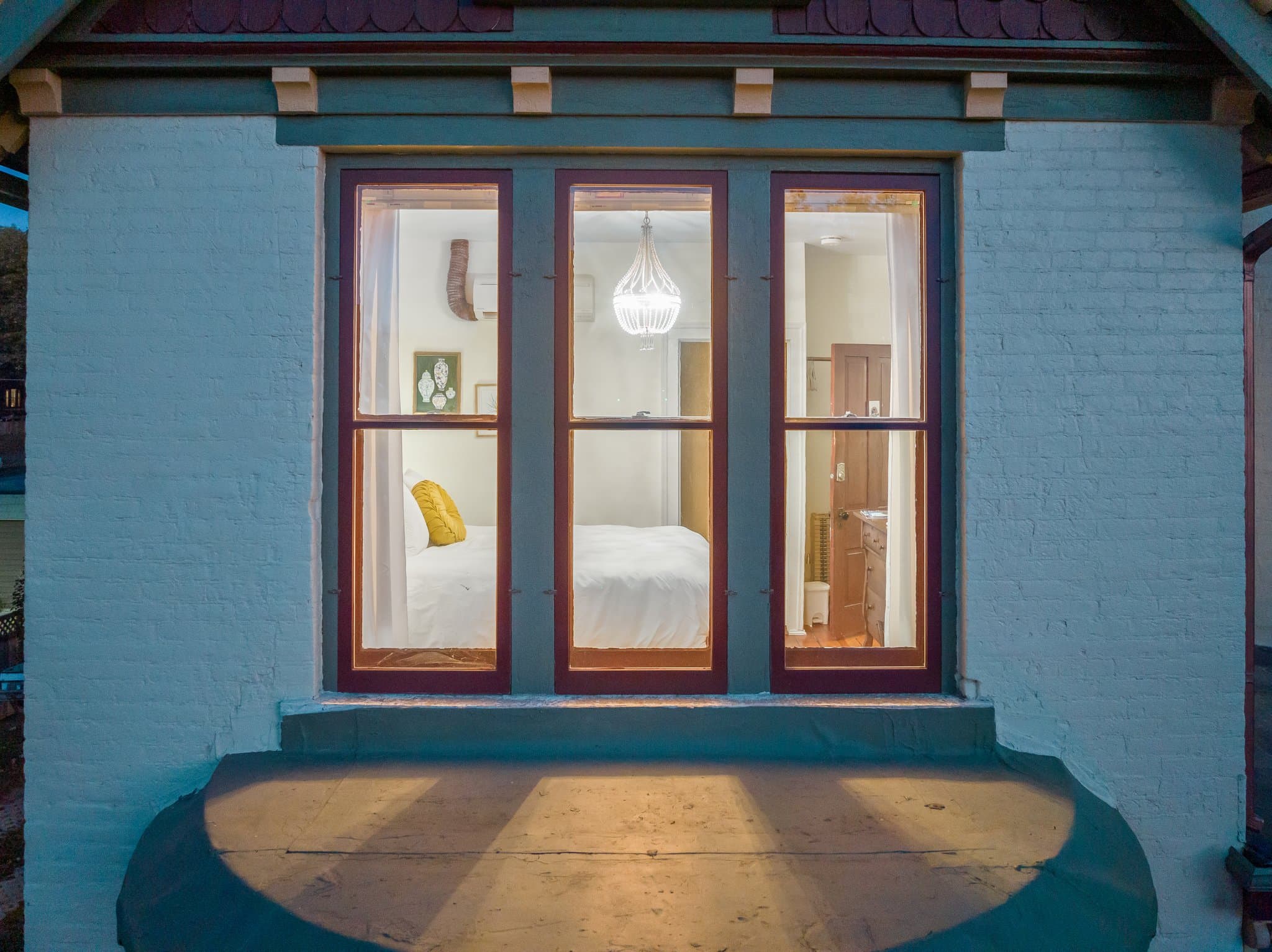 A view of a brightly lit room through a window, showcasing a cozy interior with a bed and decorative accents.